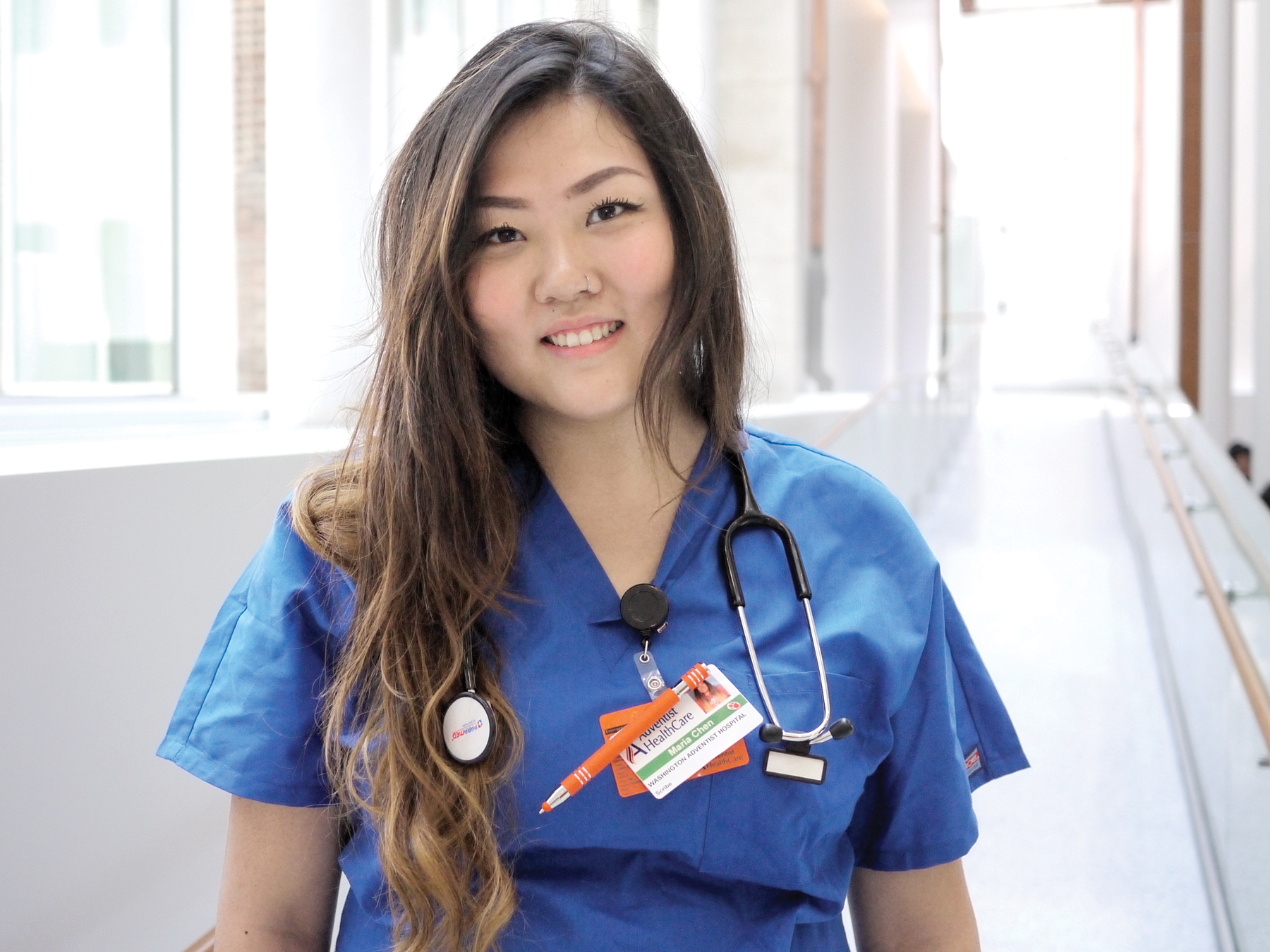 Maria Chen wearing blue scrubs and a stethoscope. She is standing at the head of a long, brightly-lit hall.