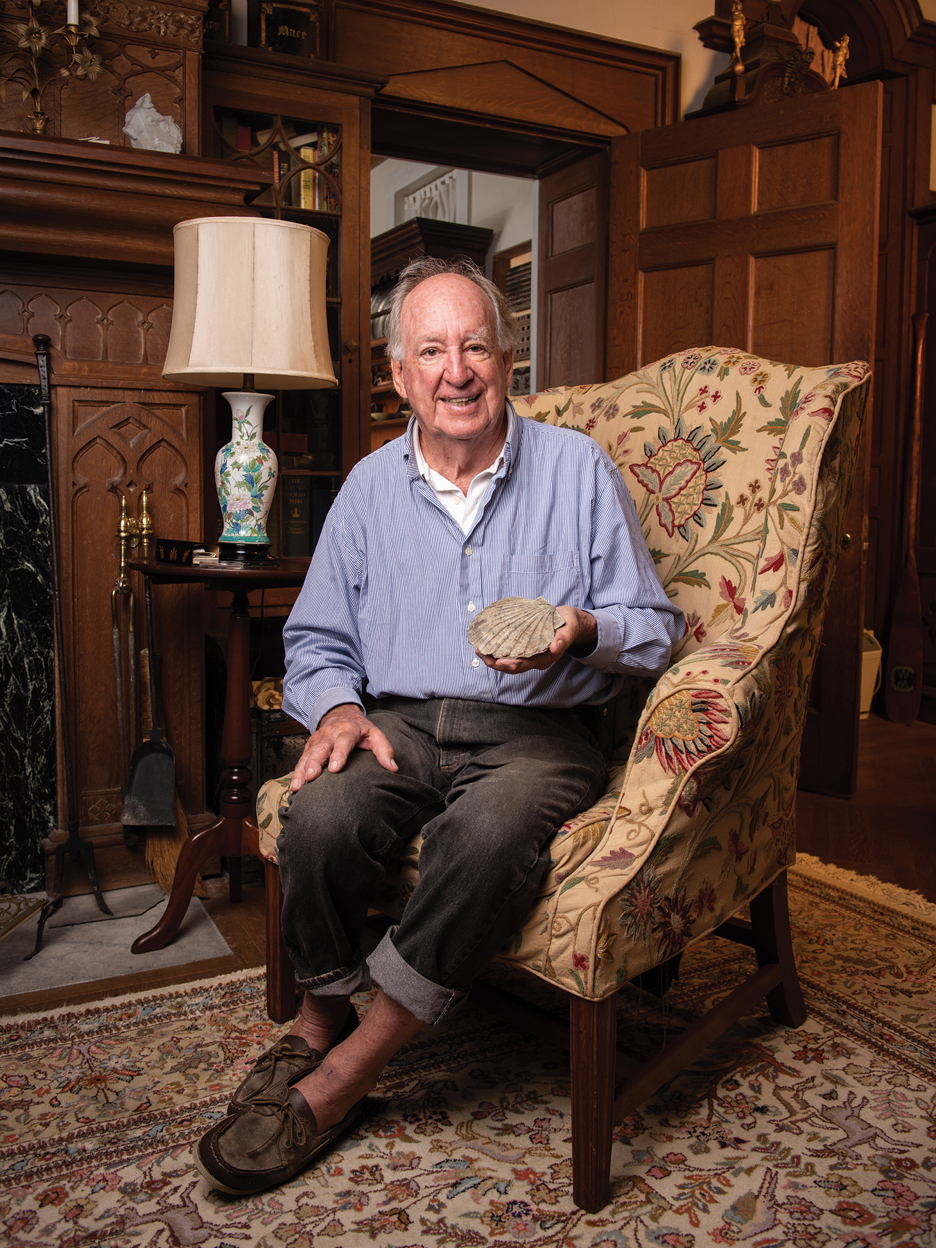 Associate Professor Emeritus of Geology Peter Stifel at the historic Hope House in Talbot County, Maryland. He is casually dressed, sitting in a chair with detailed upholstery in a room decorated with carved wooden panels, a lamp, and an elaborate rug. He is holding a large fossil clamshell.