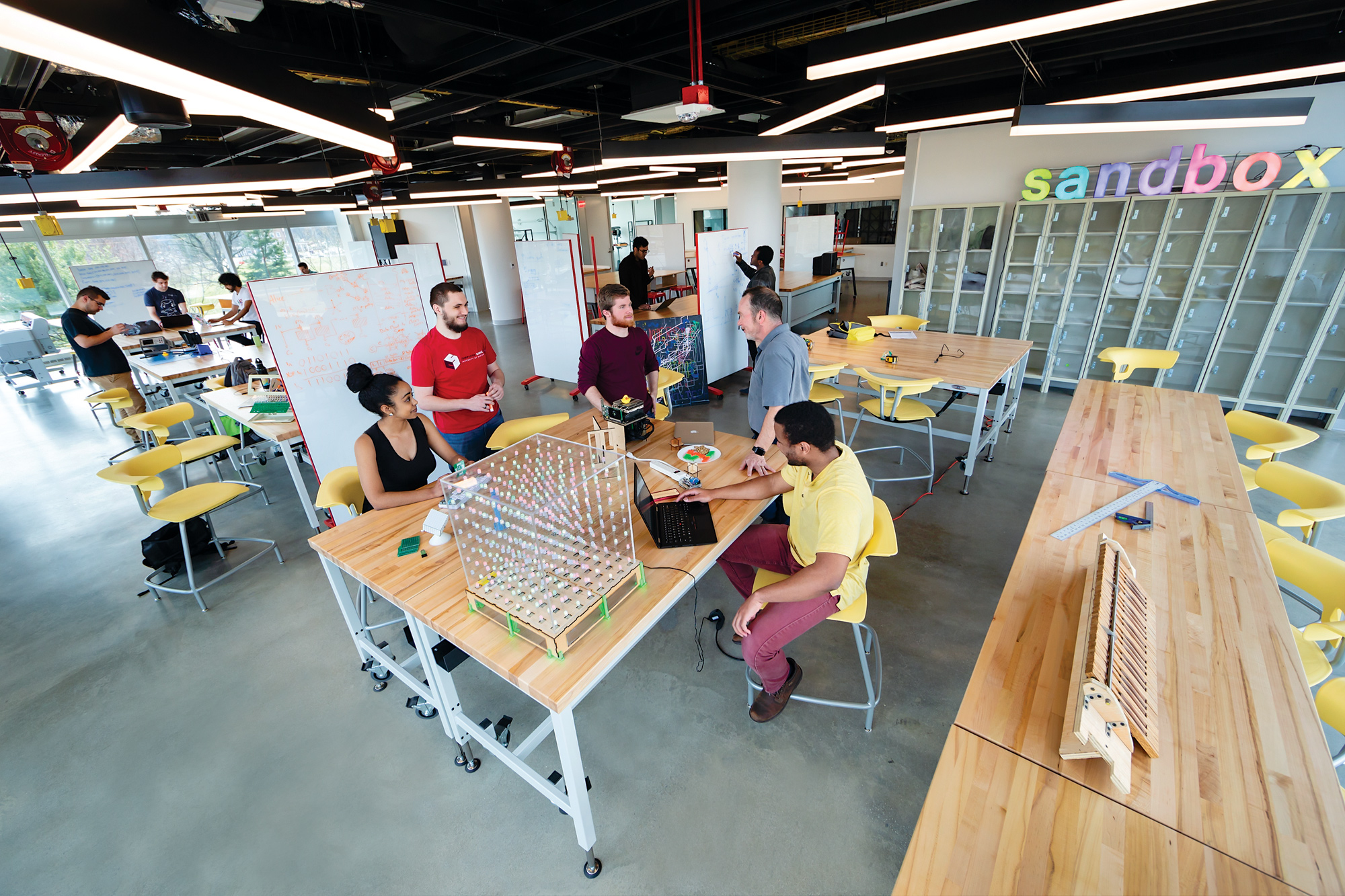 The Sandbox lab. A group of students and a professor are gathered around a table while other students work in the background. It is a very large open floorspace with a high ceiling, large windows, shelves and equipment.