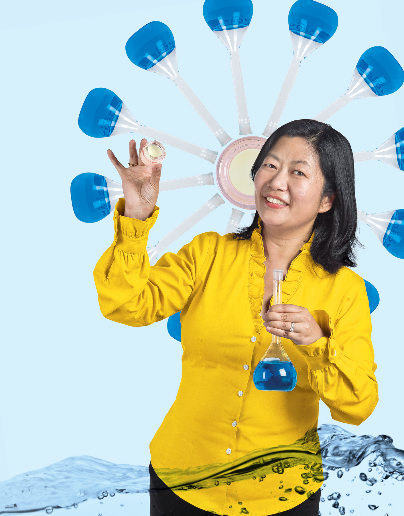 A photo illustration of Kan Cao. She is wearing a bright yellow shirt and appears to be standing in blue liquid. She is holding a flask containing blue liquid and holding a pink glass container filled with cream. Behind her, against a light blue background, is an arrangement of flasks with blue fluid in them arranged in a circle around a pink glass container, giving the impression of a flower.