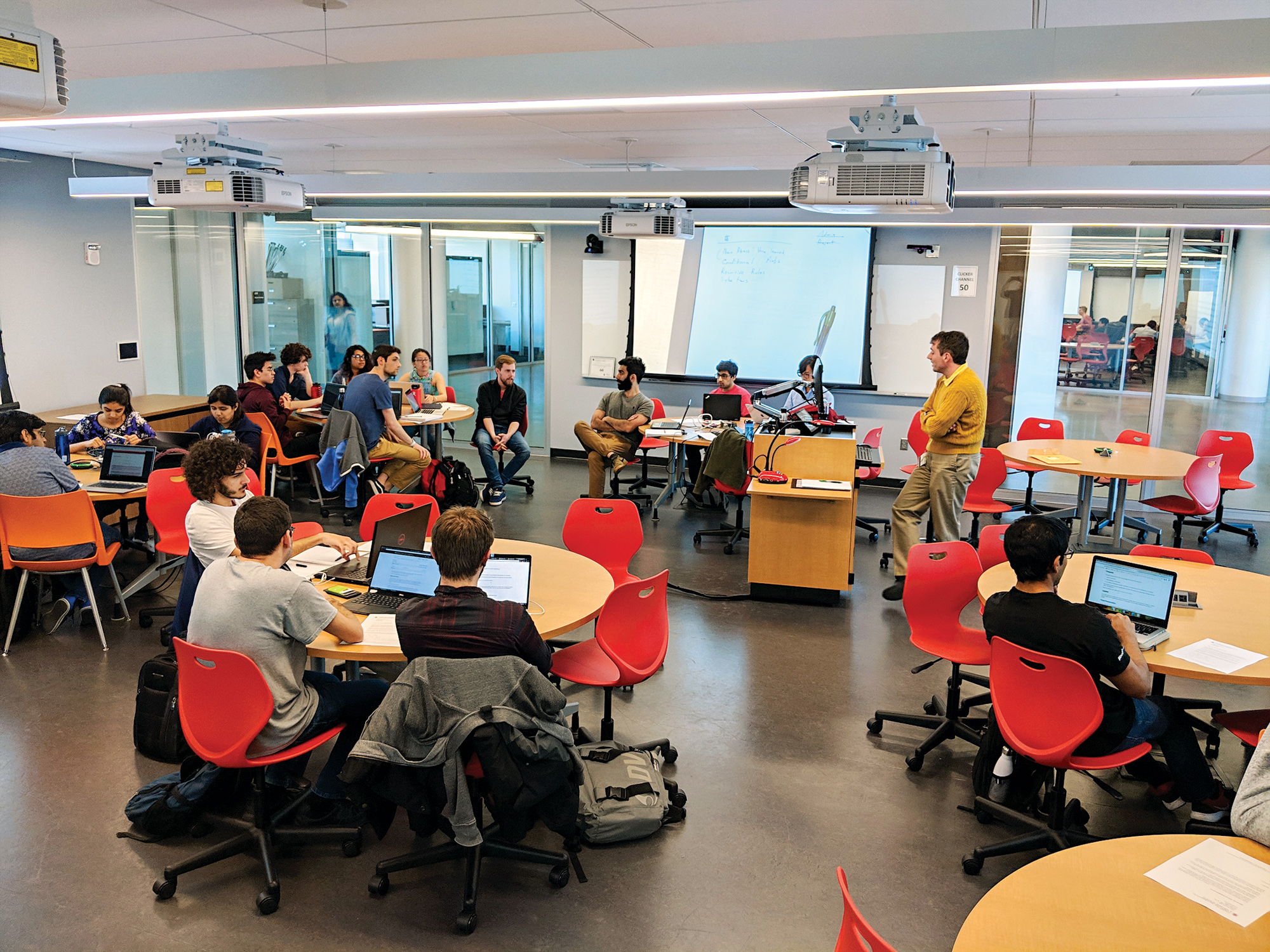 One of the Brendan Iribe Center's collaborative classrooms. It has modern styling, a glass wall and high ceilings, and students are sitting install groups at round tables.