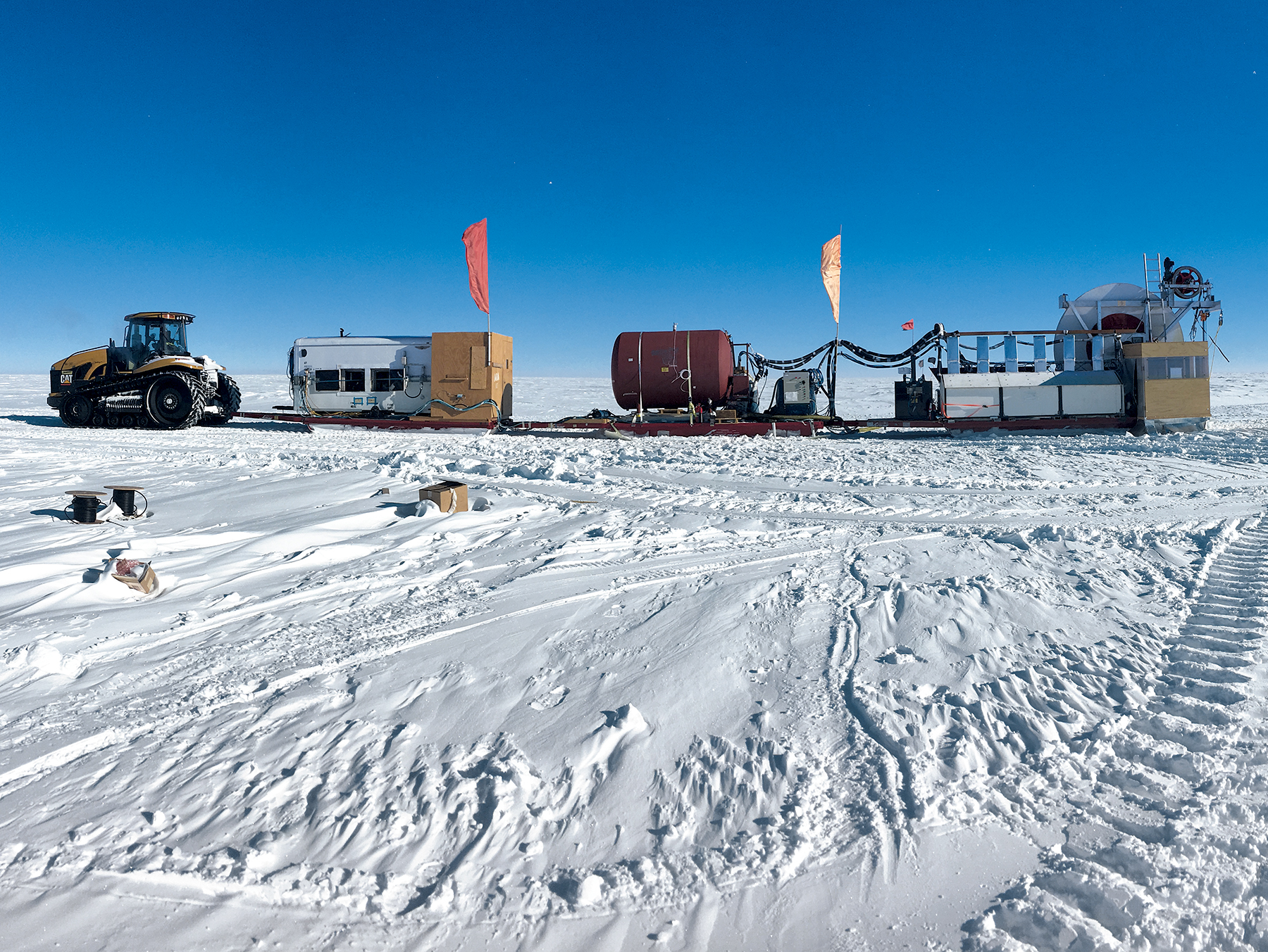 A photo of an Antarctic landscape. They sky is bright, deep blue and cloudless. The ground is covered with snow and tire tracks. A a large tractor towing scientific equipment is parked in the snow.