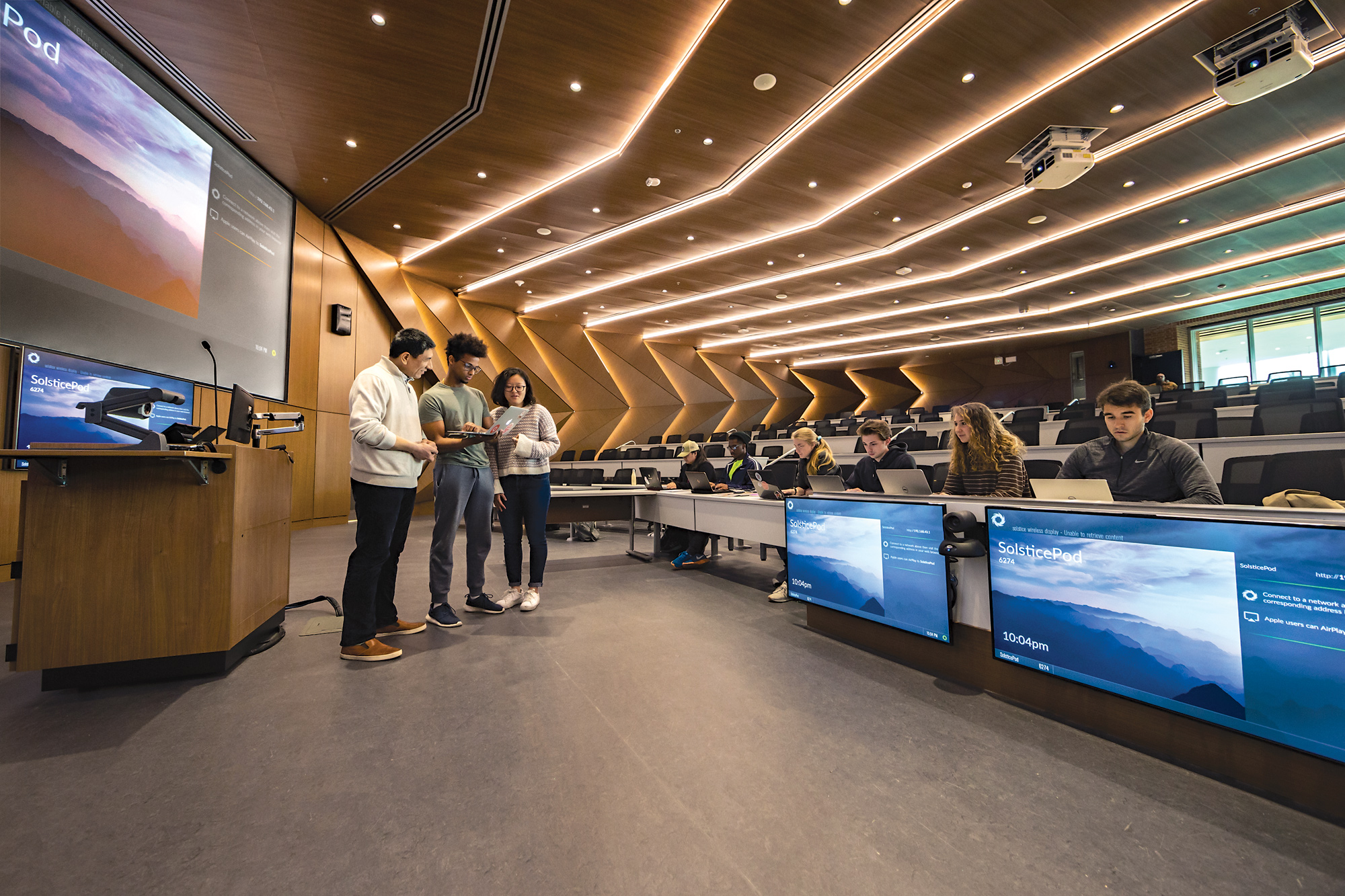 The Michael Antonov Auditorium, a wide room with multiple screens up front, tiered rows of tables, modern wood paneling on the hight ceiling and walls, and high-impact, semi-decorative track lighting. Two students are talking to a professor at the front of the room and the first row of seats is filled with students using laptops.